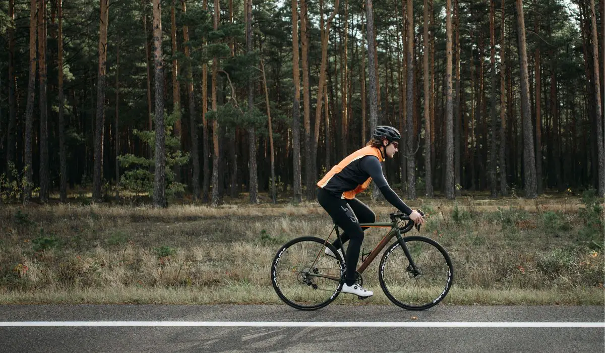 A man riding a bike in a black and orange shirt, with a black helmet on a road with forest in the background. 