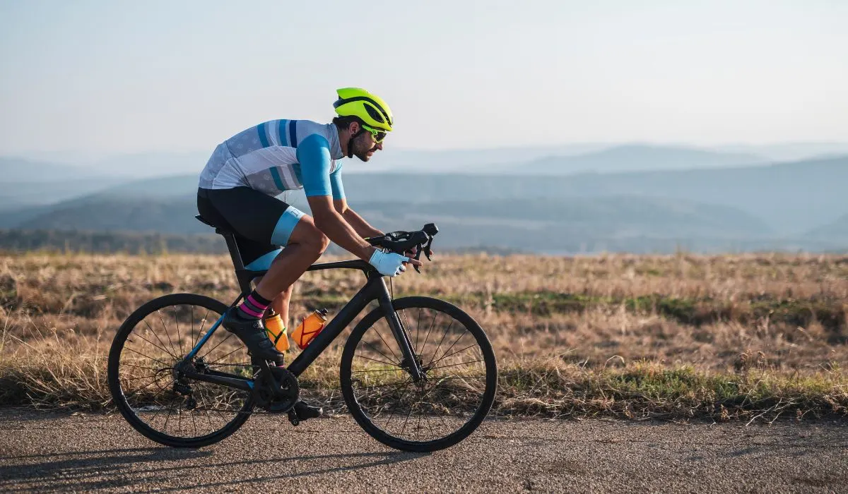 A male cyclist with a rounded hunched back, wearing a white and blue shirt and a neon yellow helmet. 