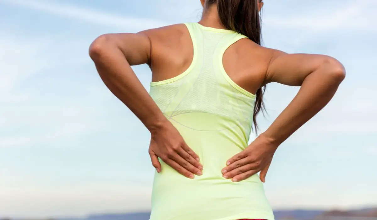 A woman with a tank top on holding her lower back in pain, back view.