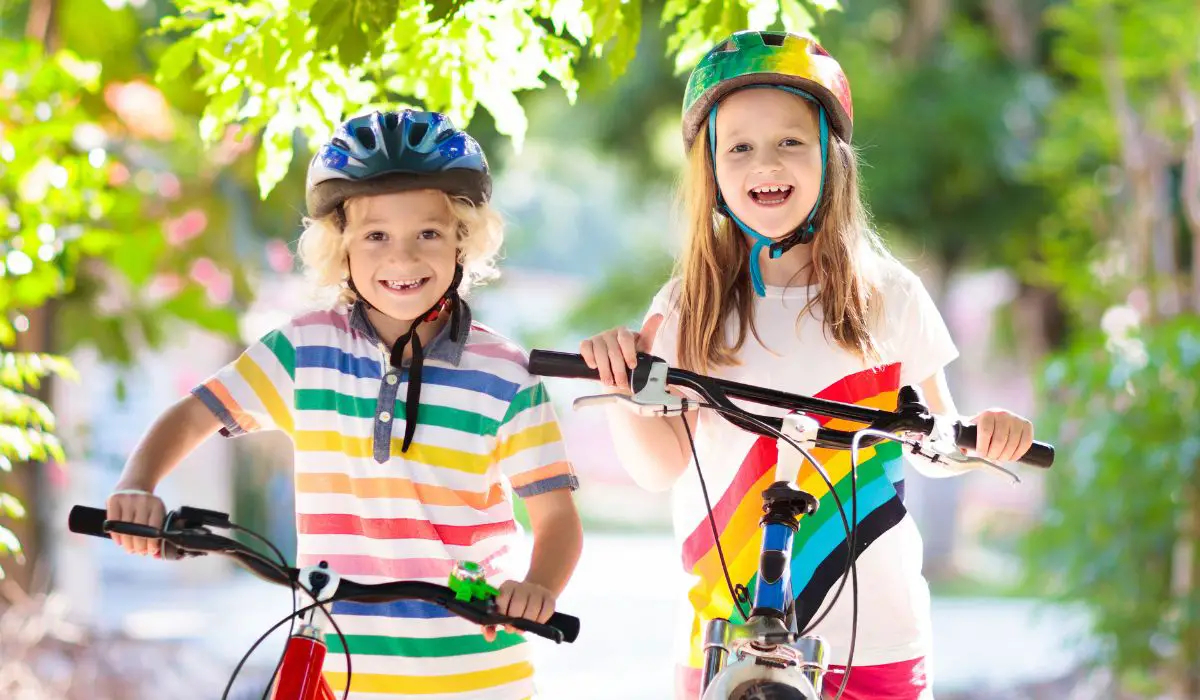 2 children riding their bikes on a sunny sidewalk surrounded by trees. 