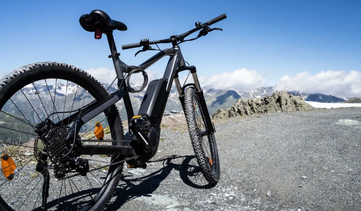 A black e-bike on a gravely mountain road with mountain tops and clouds in the background.