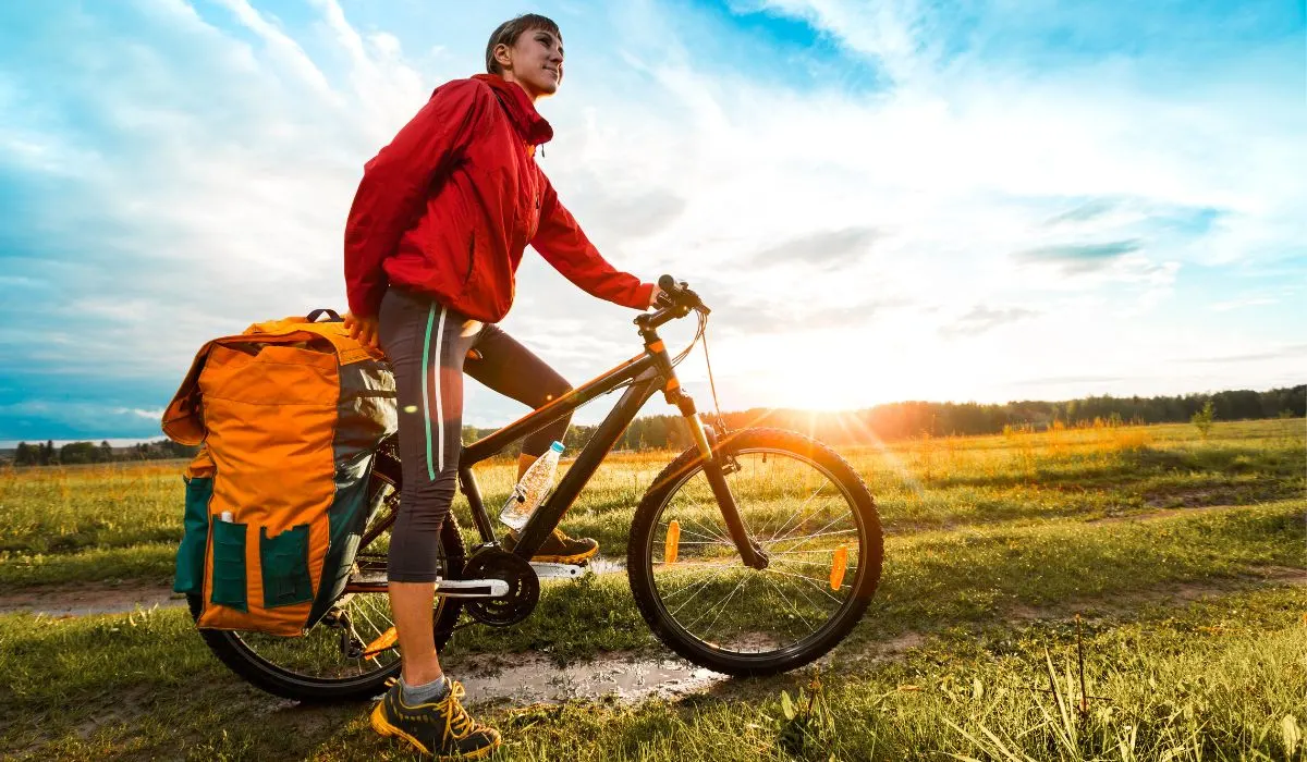 A woman on a mountain bike with a big orange pack bag on the back, on grassy terrain. 