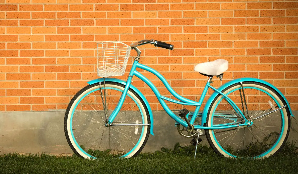 A cruiser bike that is painted turquoise, with a white basket and seat, leaning against a brick wall.