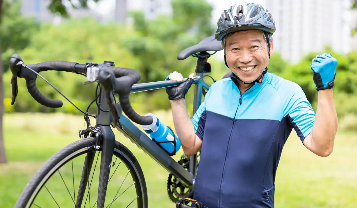 An aging person in a bike outfit holding their bike up with one hand and flexing their muscle with the other arm looking strong and happy.