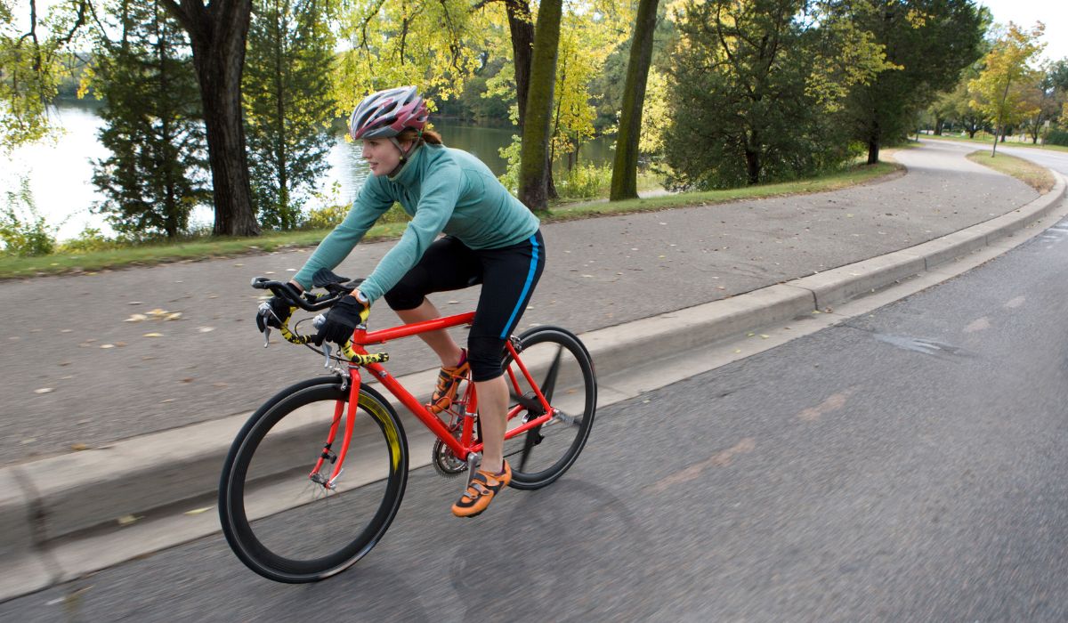 A woman on a road bike riding next to a path with green trees, side view showing proper arm reach. 