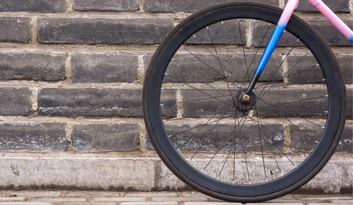 A pink and blue bike frame, photo of the front wheel against a grey brick building.