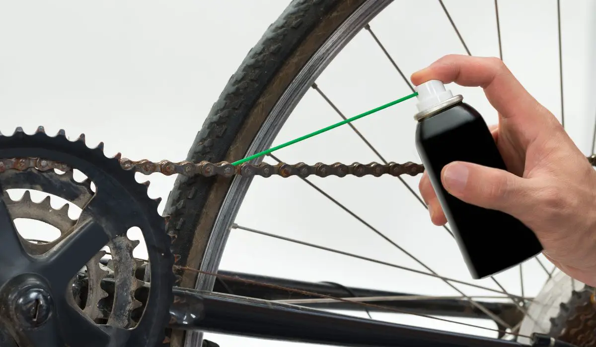 A bike chain with some rust getting lubricated with a black can that has a long green straw to aim the spray in the right spot.