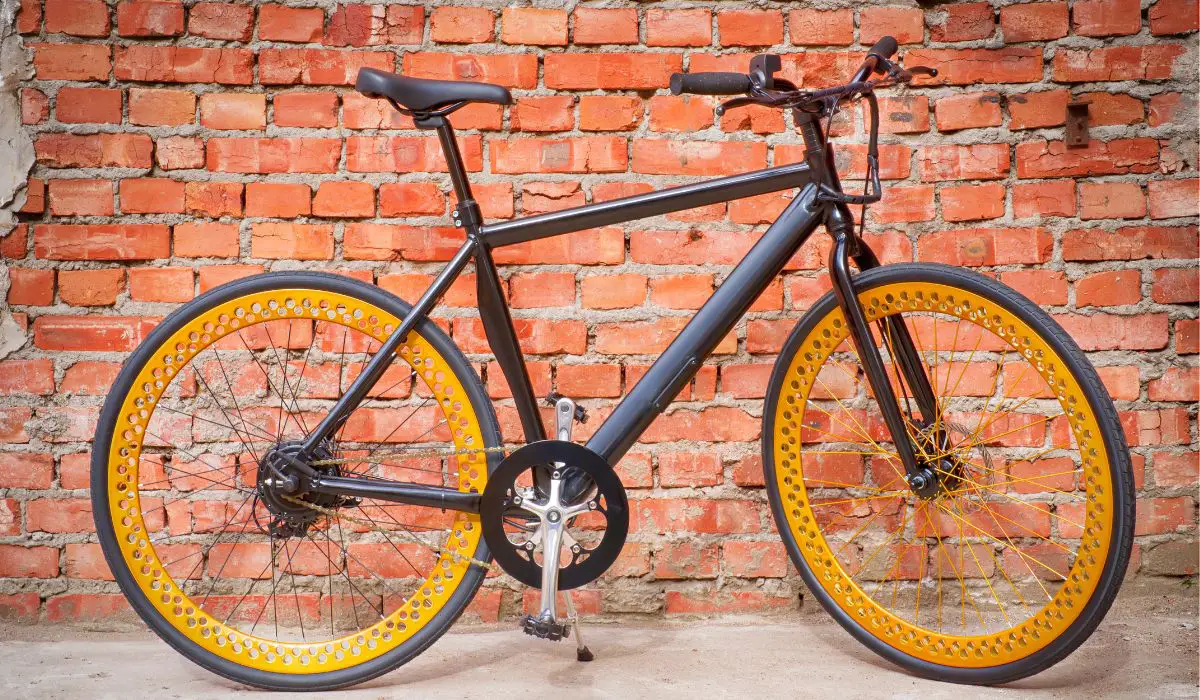 A black electric bike with orange wheels and spokes, leaning up against a brick wall. 