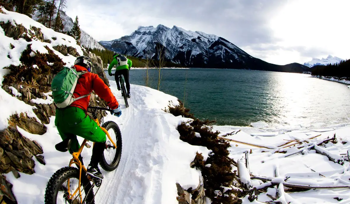 2 cyclists riding with thick tires on a snow covered road looking out at a lake. 