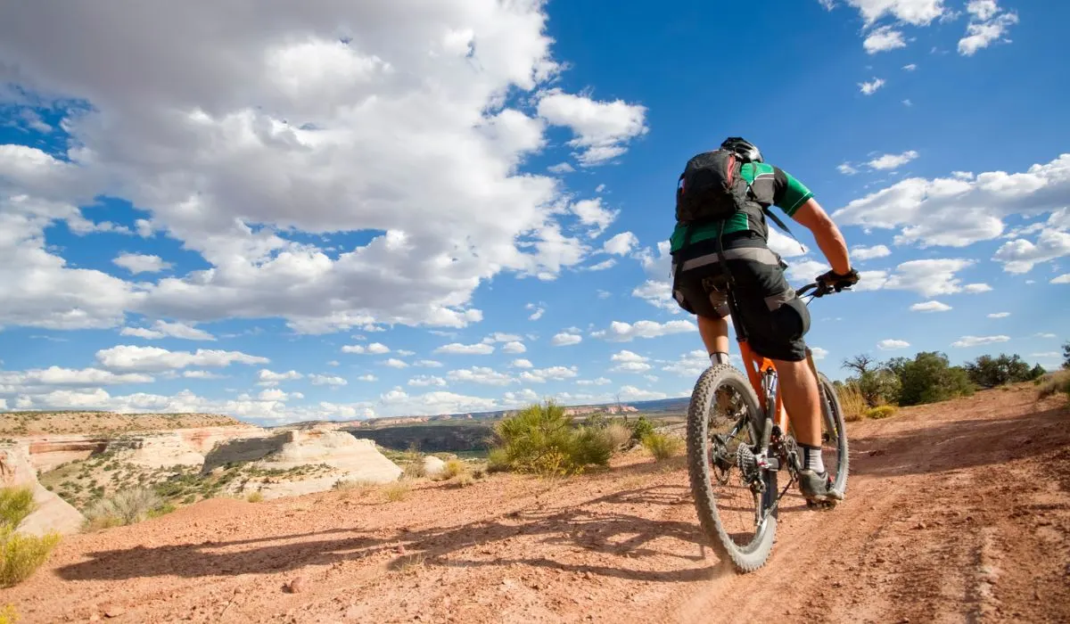 A person on a high desert mountain riding a mountain bike on a dirt path with blue sky and puffy clouds in the background. 