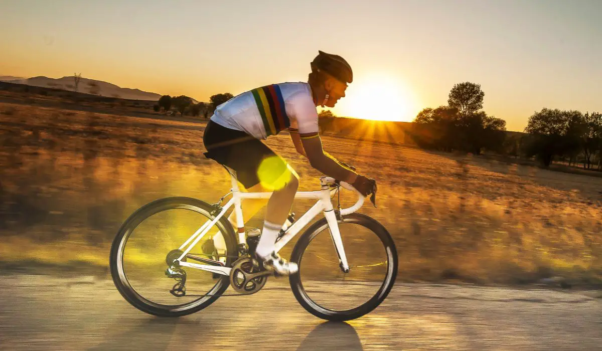 A man riding a white road bike on a country road with the sun streaming and setting behind him.