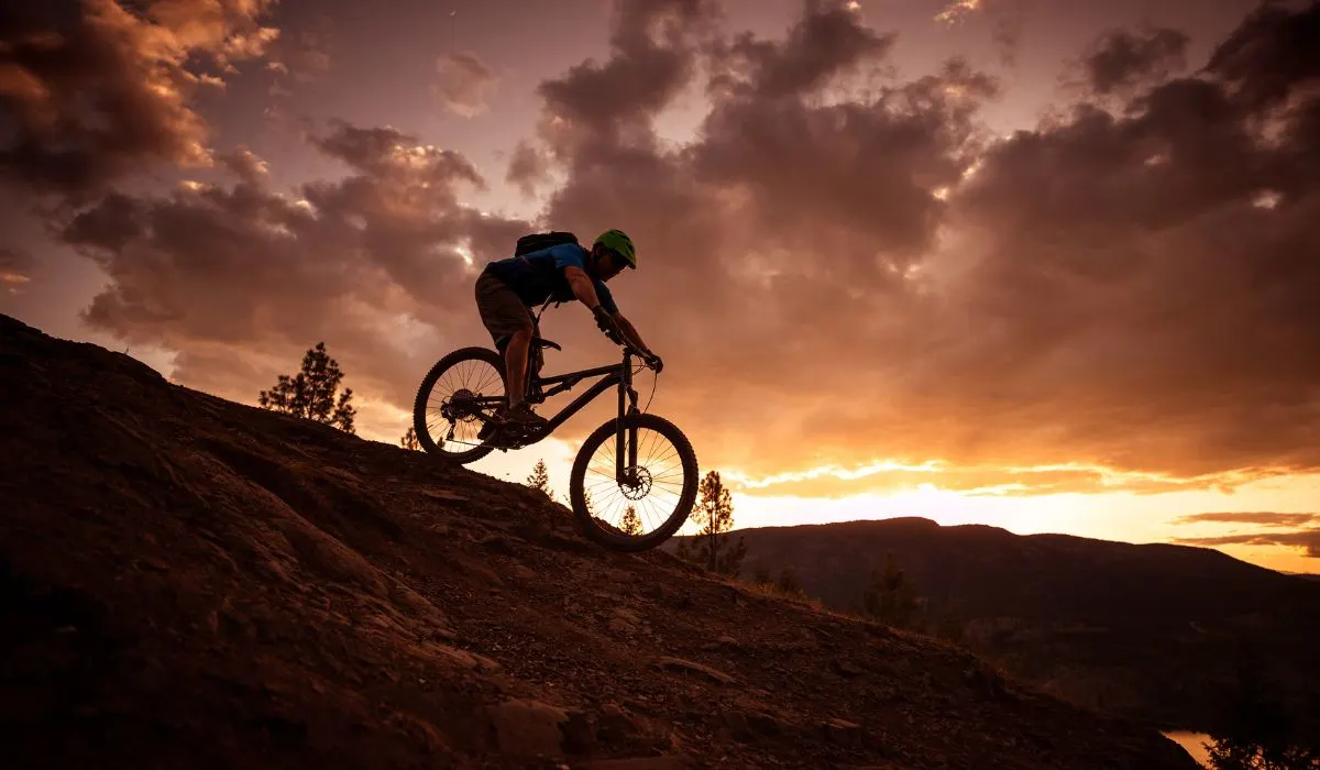 A person riding a mountain bike down a dirt hill, all is dark and there is a sunset behind.