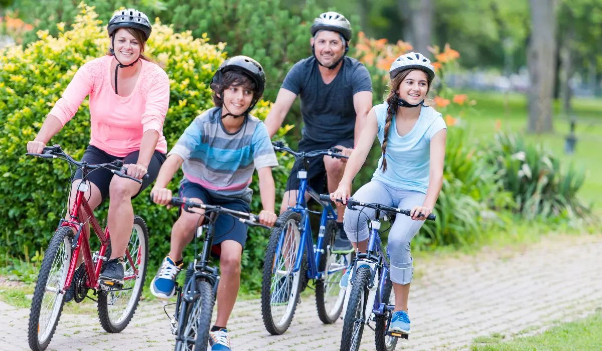 A family of 4 riding bikes on a paved path in a public park setting. 