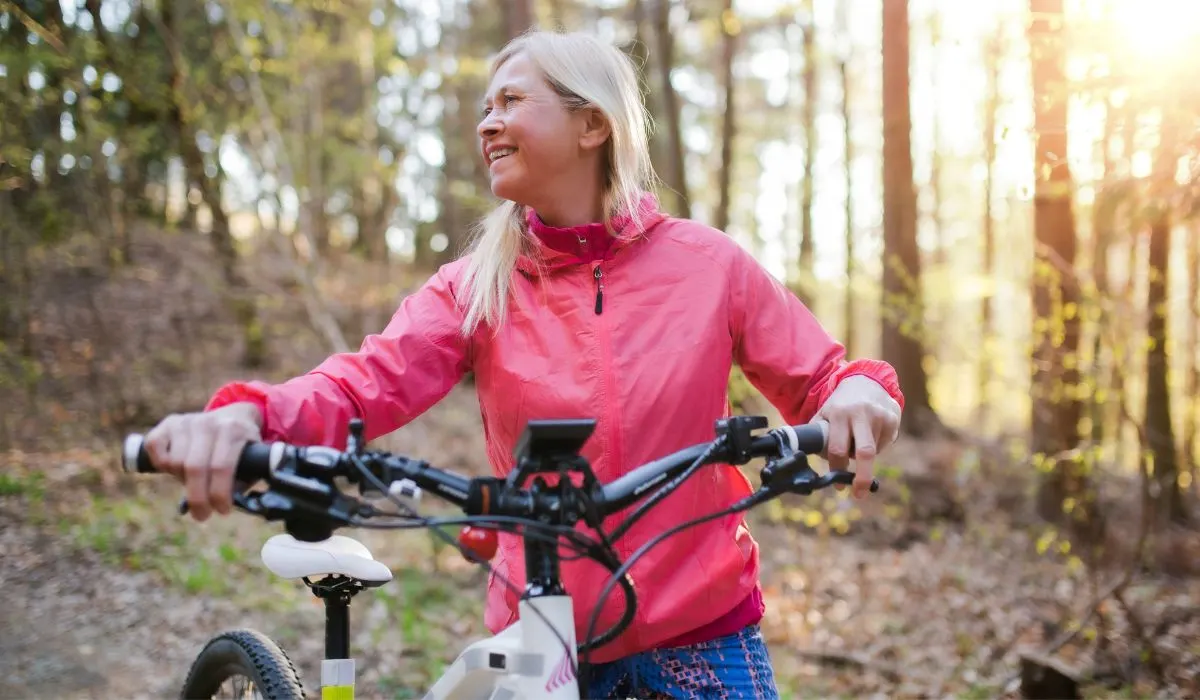 A woman with light blonde hair and a bright pink riding jacket walking her bike in the woods with sun streaming in through the trees in the background.