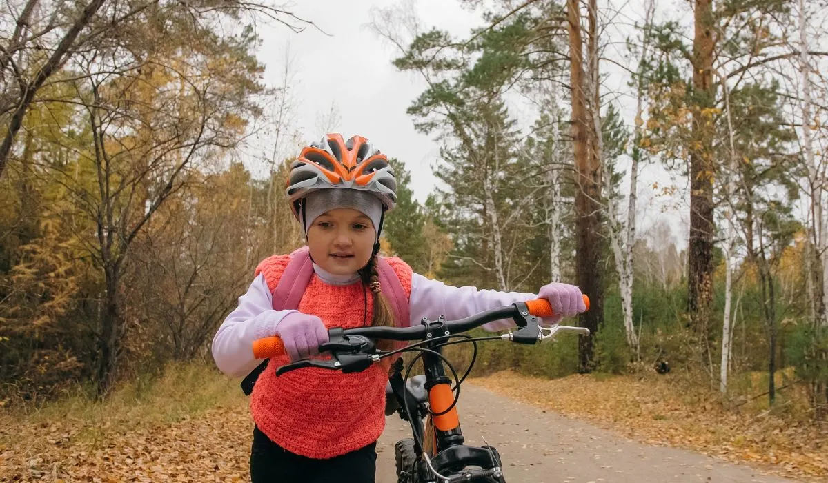 A little girl walking her bike on a wooded trail with fall leaves on the ground. 