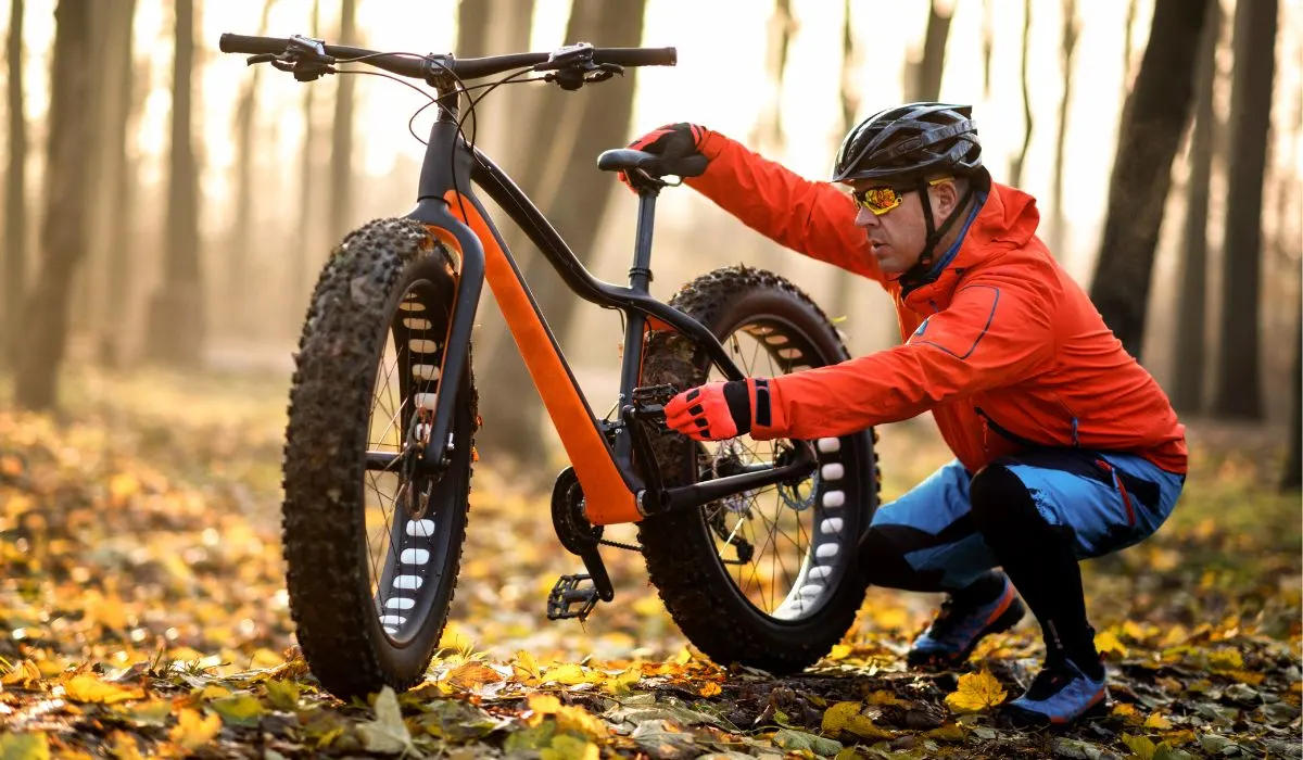 A man stopped in the woods checking the pedal on his bike. 