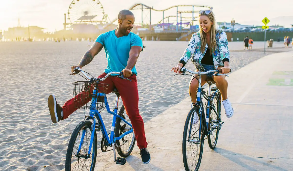 A man and woman riding bikes on the sidewalk right next to a sandy beach with the boardwalk and sun in the background in Santa Cruz, CA. 