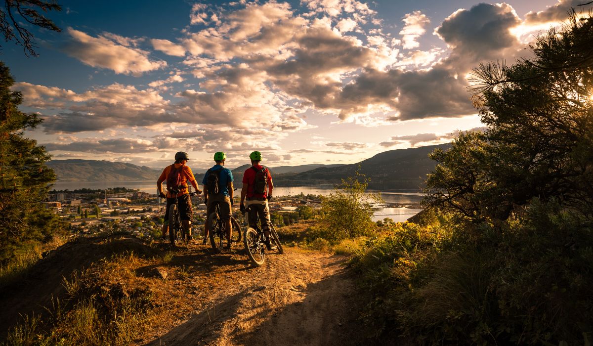 3 mountain bike cyclists on a trail at the top of a hill, overlooking a town and a river with a sunset.