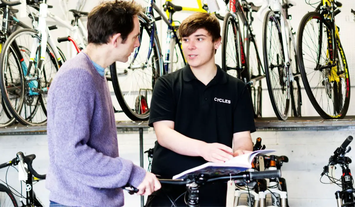 A man working at a bike shop with explaining to another man about bikes with a book open for reference and bikes for sale in the background.