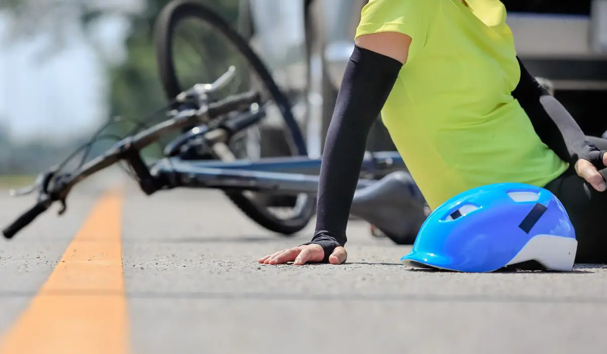 A person sitting on the road with their helmet off in front view, and a crashed bike with a car behind it in the background. 