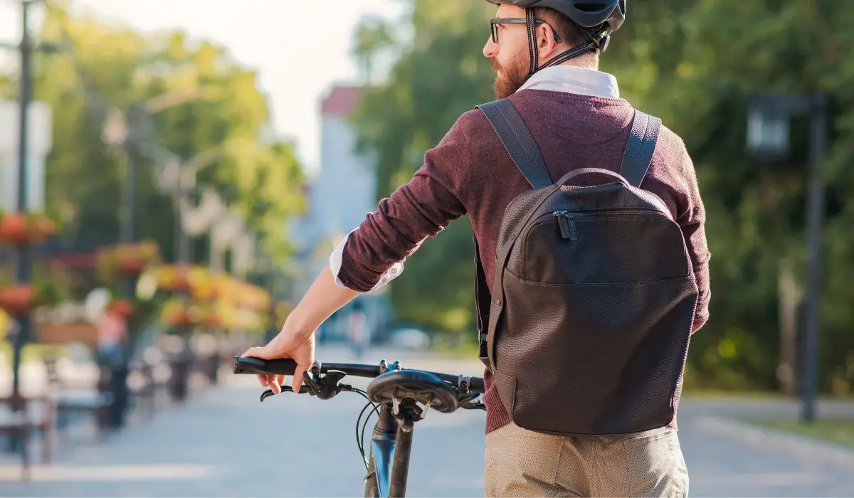 A man in casual work clothes with a backpack and helmet walking his bike. 