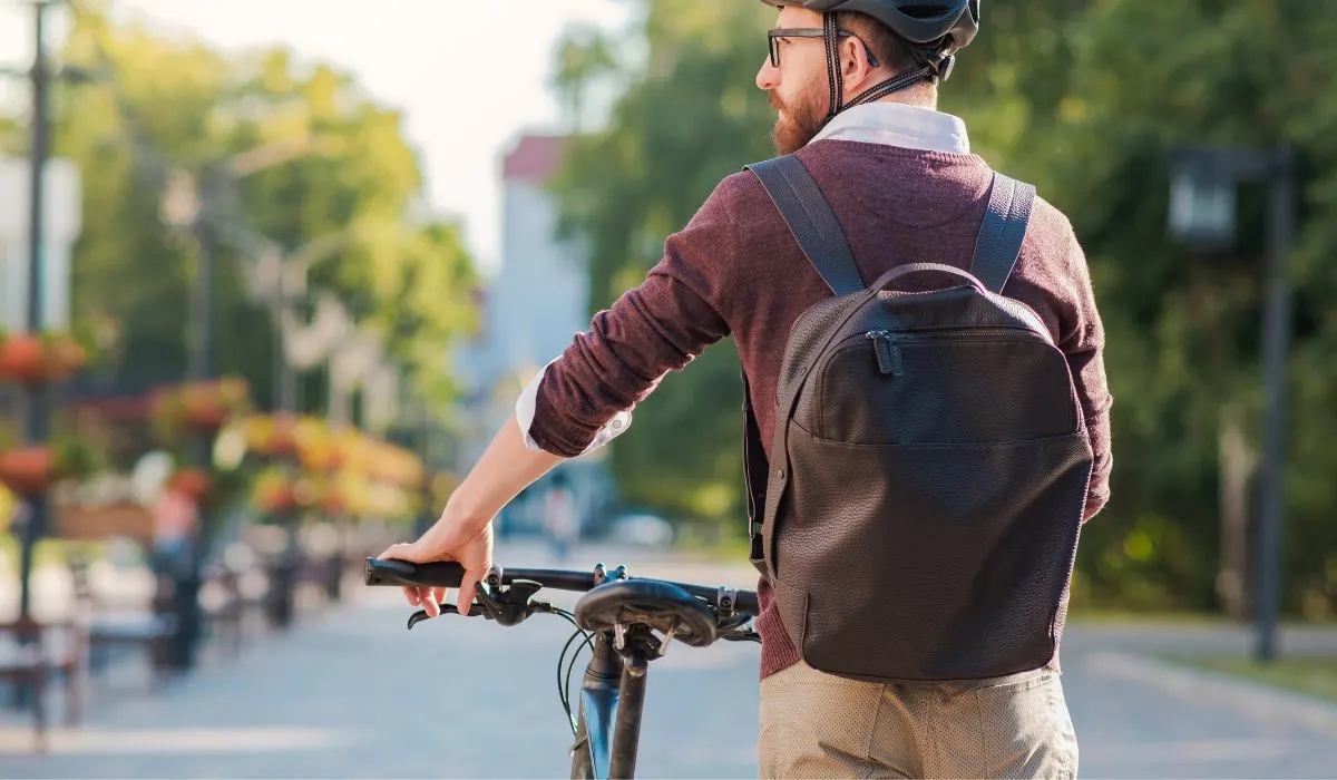 A man in casual work clothes with a backpack and helmet walking his bike.
