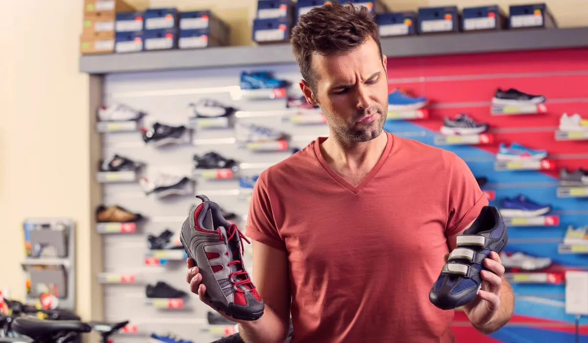 A man in a store with different cycling shoes in each hand with a perplexed look on his face.  
