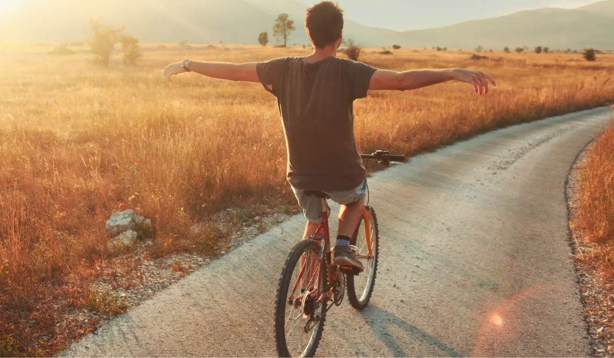 A man riding a bike on a country road with his arms out feeling freedom, with a sunset. 