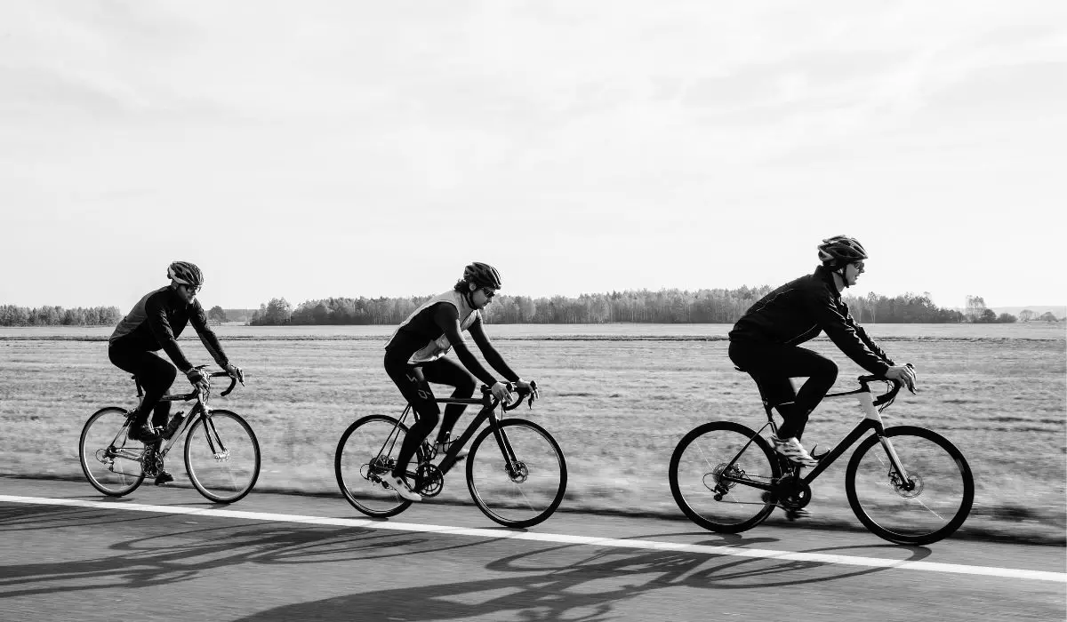 A black and white photo of 3 people riding bikes in a row, side view. 