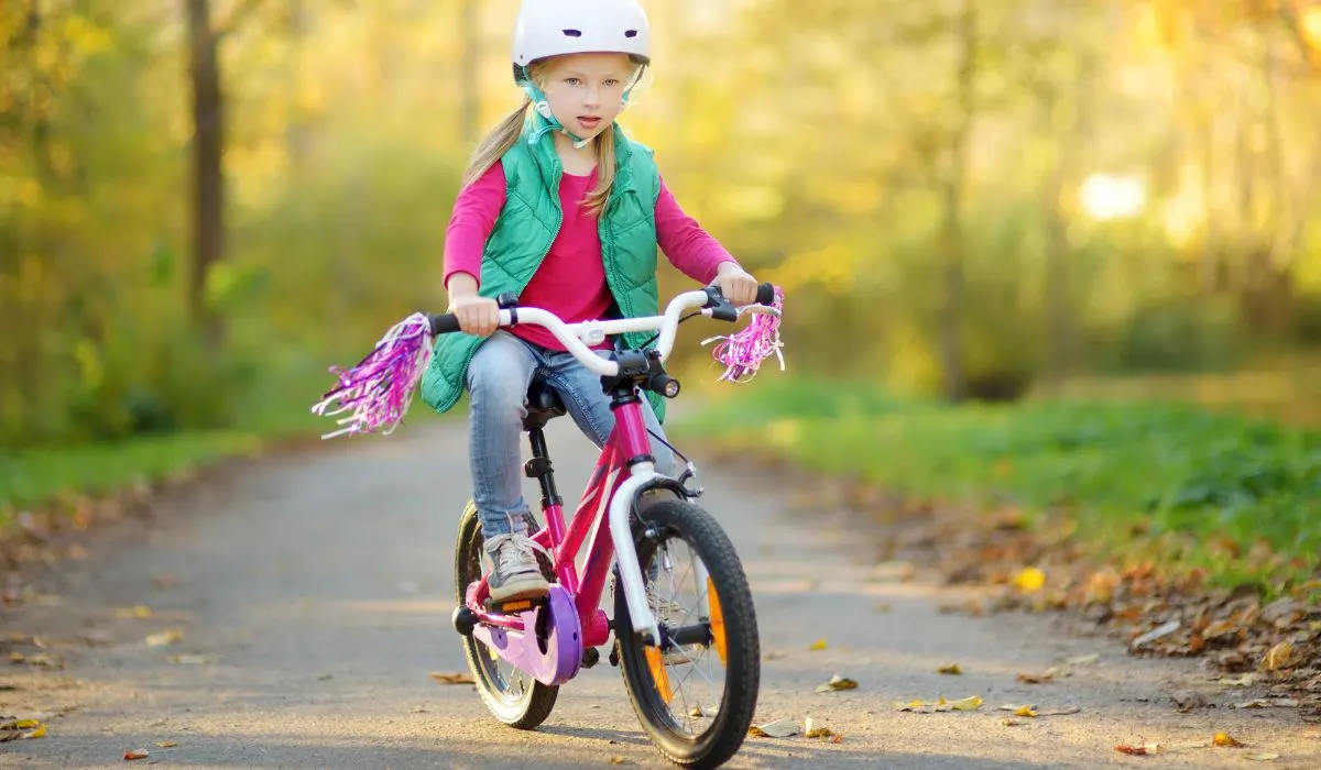 A little girl with blonde ponytails riding a pink bike with pom poms on the handlebars. 