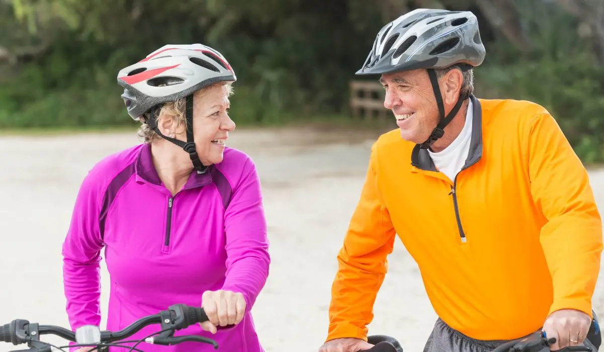 An aged couple pushing bikes and smiling at each other, both wearing helmets. 