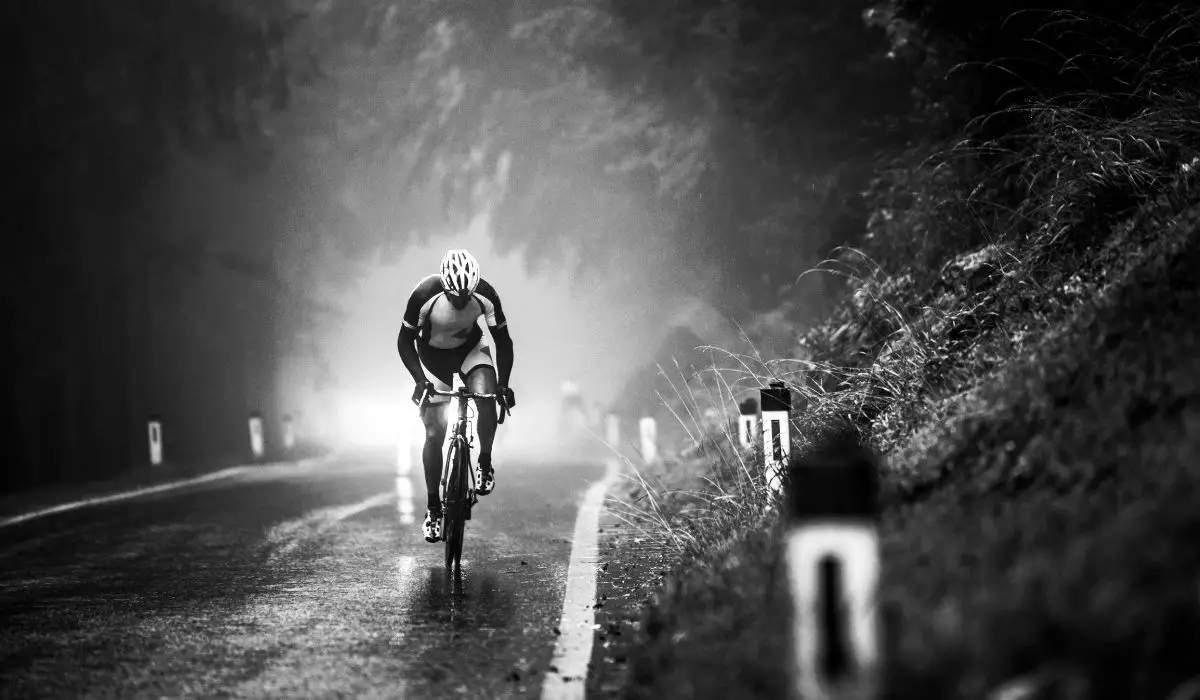 A black and white photo of a road cyclist riding on a paved forested road, front view.
