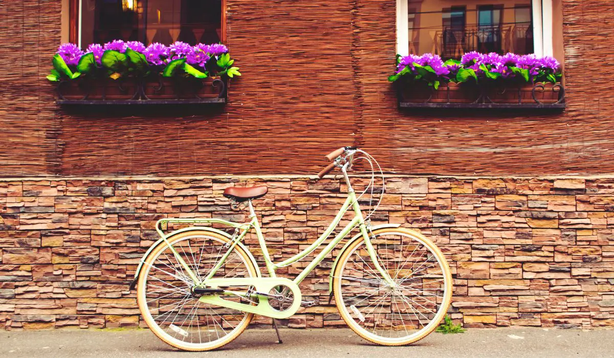 A retro bike in lime green leaning against a brick wall outside, with window ledges that have purple blooming flowers on them. 