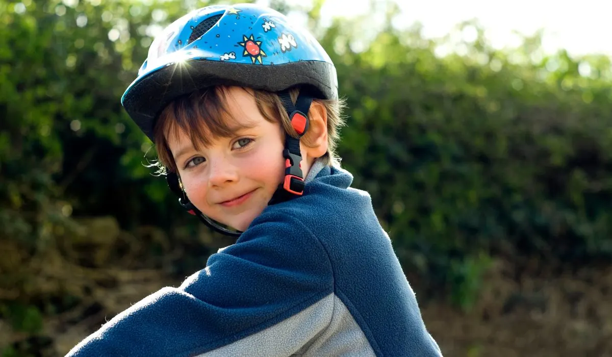 A little boy with a bike helmet on reaching out to his bike handlebars and turning his head to the side to face the camera.