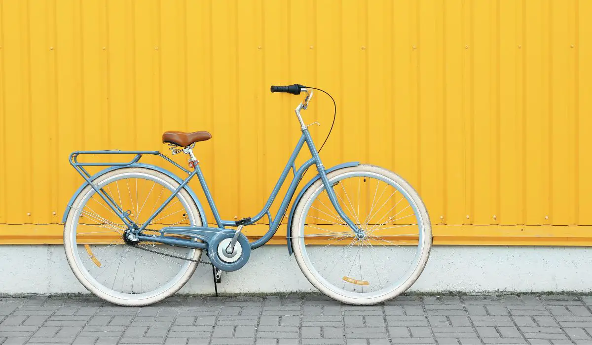 A blue retro style cruiser bike leaning against a yellow wall on a grey brick paver ground. 