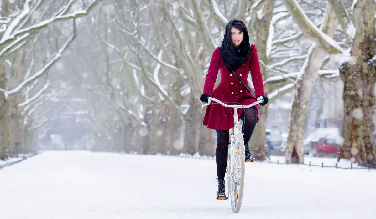 A woman in a red coat with gold buttons and a black scarf over her hair riding a bike on a snowy road.