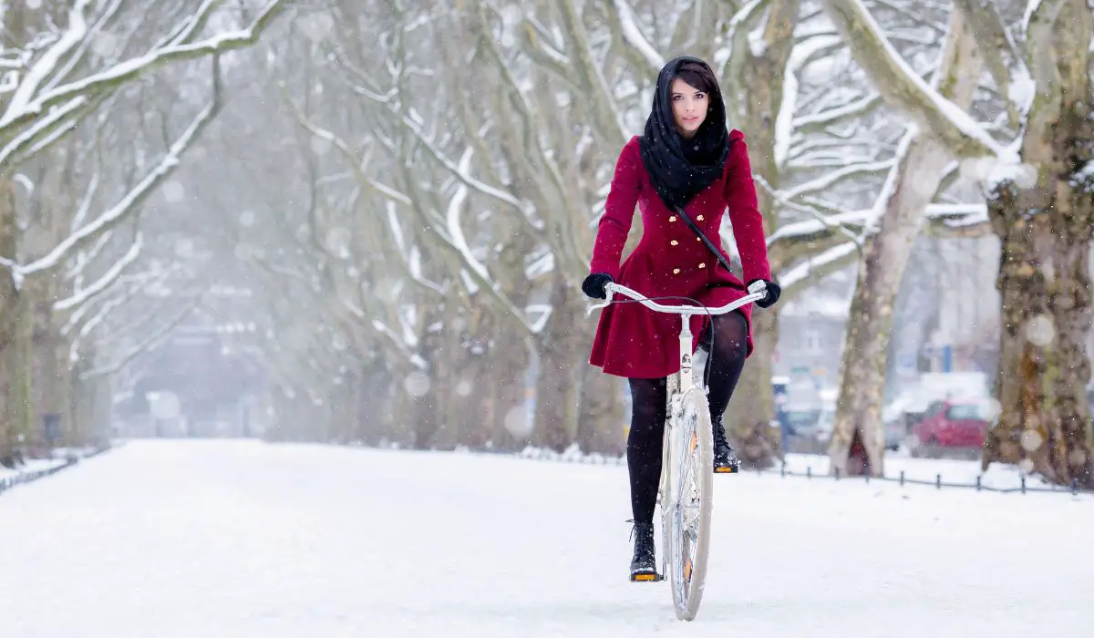 A woman in a red coat with gold buttons and a black scarf over her hair riding a bike on a snowy road. 